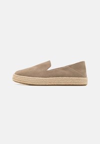 Beige Wildlederschuh mit einer geflochtenen Jutesohle, der eine gebogene Oberseite, ausgeschnittene Seiten und ein flaches Profil aufweist.