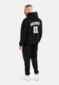 Felpa nera in cotone con "SMILODDY 01" stampato in bianco sulla schiena, abbinata a pantaloni neri coordinati e sneakers bianche.