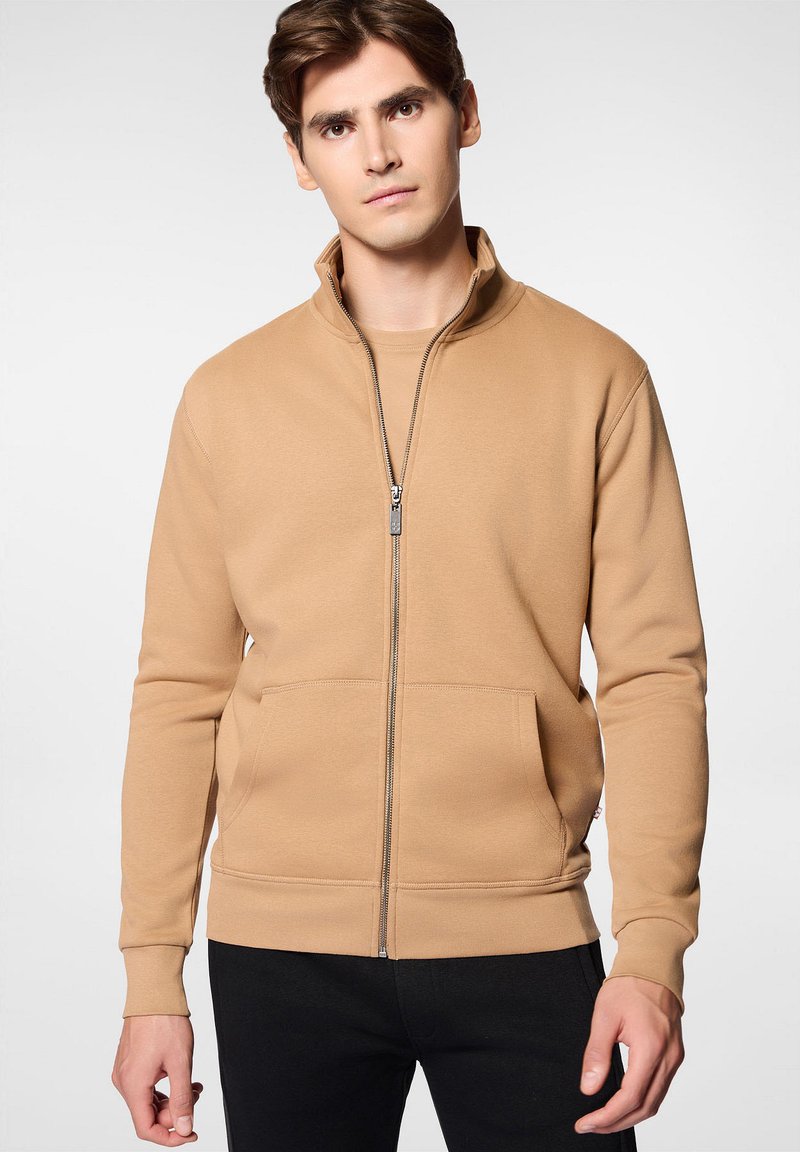 Khaki sweter rozpinany z wysokim kołnierzem, przednimi kieszeniami i ściągaczami na rękawach. Miękka, gładka tekstura z dopasowanym krojem; noszony na dopasowanej bluzce.
