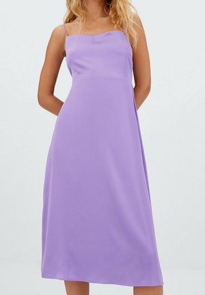 Femme portant une robe midi sans manches, de couleur violet clair, avec un décolleté carré et de fines bretelles, sur un fond uni.