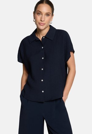 Femme portant une chemise à manches courtes texturée bleu marine avec col et boutons, associée à un pantalon bleu marine, debout les mains dans les poches.