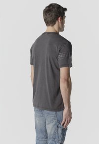 Homme vu de dos portant un T-shirt à manches courtes gris foncé et un jean cargo bleu clair, sur un fond clair uni.