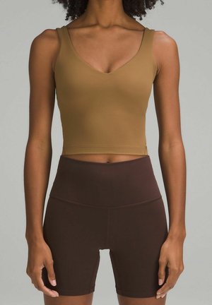 Mujer con un top corto marrón y pantalones cortos de ciclista marrón oscuro de cintura alta, de pie frente a un fondo liso.