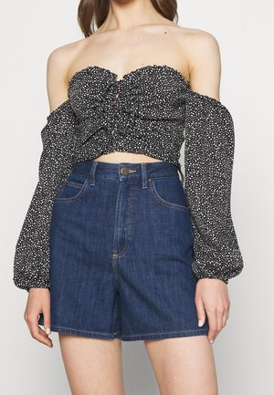 Kvinde iført høj talje mørkeblå denimshorts og en sort crop top med lange ærmer og hvide pletter, med rynkedetalje foran.