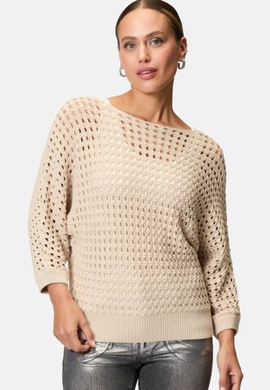 Beige Strickpullover mit einem offenen Maschenmuster, Boudekolleté und 3/4-Ärmeln. Rippbündchen und Saum. Kombiniert mit glänzenden, metallischen Slim-Fit-Hosen.