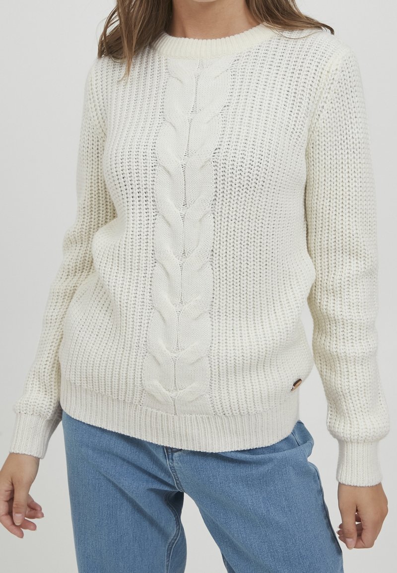 Femme portant un pull en maille torsadée crème avec poignets et ourlet côtelés, associé à un jean bleu clair, montré du cou aux hanches.