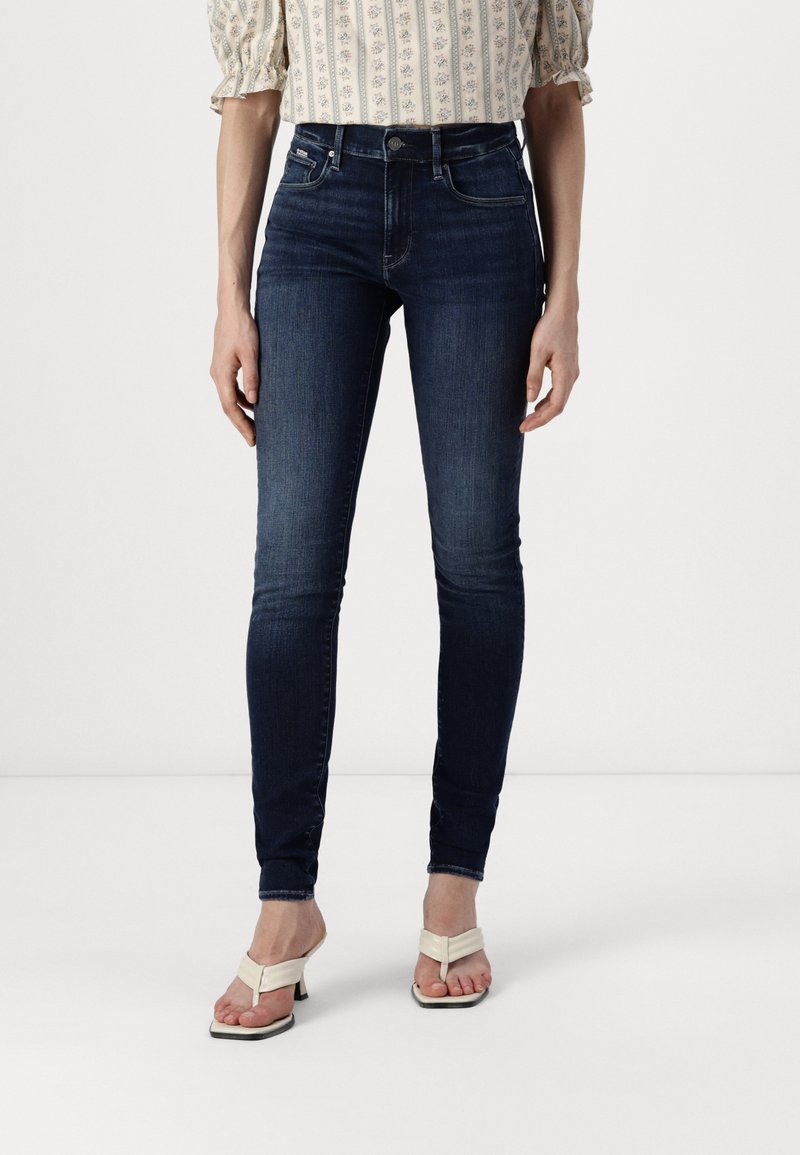 Mörkblå skinny jeans gjorda av denim. Har hög midja, slim fit och minimalistisk hårdvara. Matchas med ljusa sandaler.