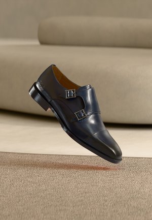 Scarpa elegante doppia fibbia monk in pelle nera sospesa sopra un tappeto beige, con un divano beige chiaro sfocato sullo sfondo.