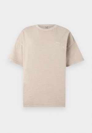 T-shirt en coton beige avec des manches courtes, col rond, et un petit texte imprimé blanc "NÉ AU PARADIS" sur la poitrine. Texture lisse.