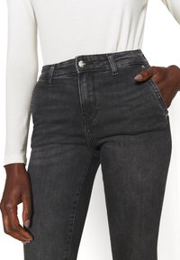 Jeans de denim negro con una cintura alta, un ligero desgaste, bolsillos frontales y cierre de botón metálico. Detalle de costura a lo largo de las piernas.