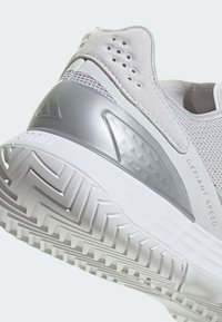 Hellgrauer und weißer Sportschuh mit strukturierter Sohle, Wildlederferse, Mesh-Obermaterial, silberner Fersenstütze und „DEFIANT SPEED“ seitlich aufgedruckt.