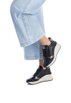 Las zapatillas negras y bronce presentan un diseño acolchado, acentos suaves y una suela blanca gruesa. Combinadas con unos pantalones vaqueros cortados de color azul claro.