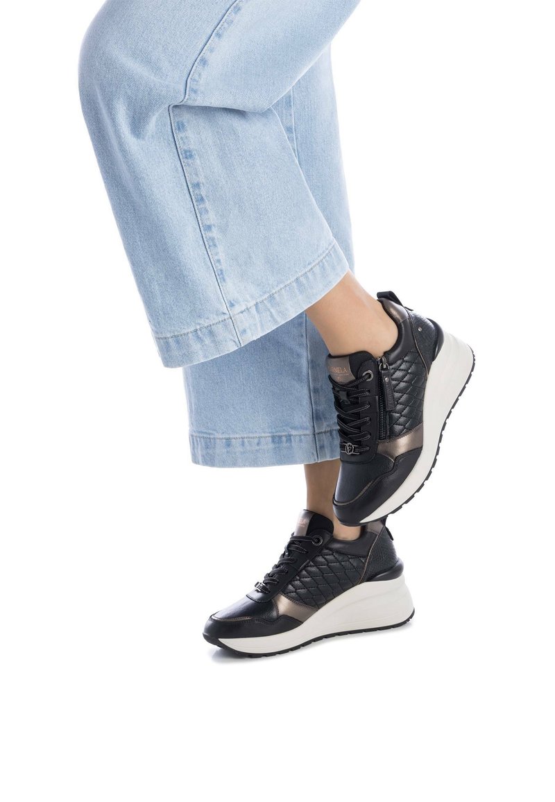 Las zapatillas negras y bronce presentan un diseño acolchado, acentos suaves y una suela blanca gruesa. Combinadas con unos pantalones vaqueros cortados de color azul claro.