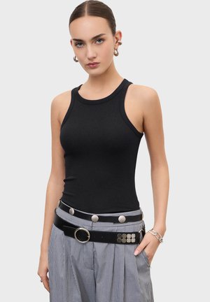 Mladá žena s uhladenými vlasmi, oblečená v čiernom bezrukávovom rebrovanom topu a károvaných nohaviciach s čiernym dvojitým opaskom a striebornými doplnkami.