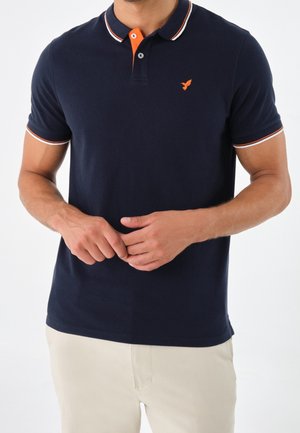 Mand iført en marineblå polo med krave og ærmekanter i orange og hvide striber samt beige bukser, hænder let foldet.