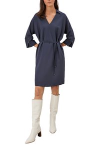 Donkerblauwe jurk met een V-hals en driekwart mouwen, met een ceintuur op de taille, gecombineerd met witte leren laarzen tot de knie. Zachte textuur.