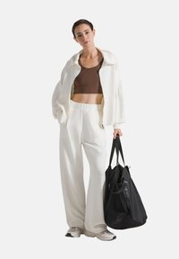 Weiße, übergroße Jacke über einem braunen Cropped-Top, kombiniert mit lockeren weißen Hosen. Schwarze Tasche mit kurzen Griffen. Helle Sneaker.