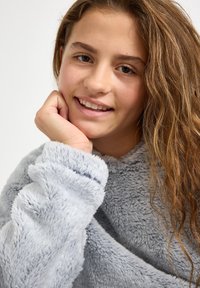 Hellgrauer, weicher, flauschiger Kapuzenpullover aus Stoff mit weitem Kragen und Bündchen an den Ärmeln. Das Modell hat langes, welliges Haar und stützt ihr Kinn auf die Hand.