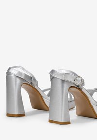 Sandalias de tacón alto plateadas con un tacón cuadrado, hechas de material satinado. Presentan correas entrelazadas y un cierre de hebilla. La suela es de color marrón claro.