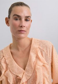 Mujer con el cabello rubio recogido lleva una blusa melocotón con volantes y un sutil patrón de leopardo, contra un fondo claro y simple.