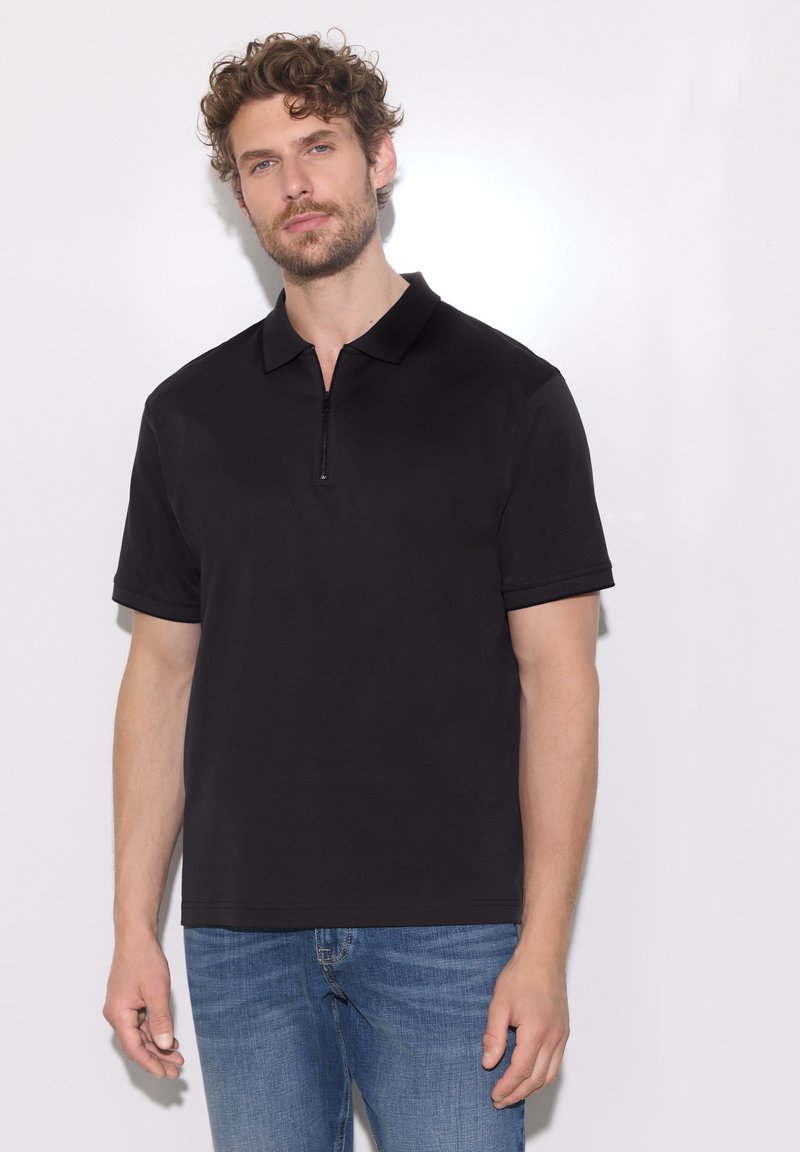 Homme aux cheveux bouclés portant un polo noir à manches courtes et un jean bleu, debout devant un fond blanc uni.