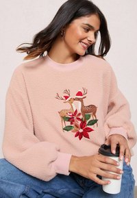 Maglione in pile rosa con maniche lunghe, caratterizzato da un ricamo di renne e un design floreale. Il modello tiene una tazza di caffè.
