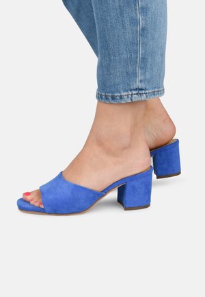 Niebieskie zamszowe klapki na obcasie z szerokim, otwartym przodem, grubym obcasem i gładką fakturą. Połączone z jasnymi jeansami, odsłaniającymi pedicure.