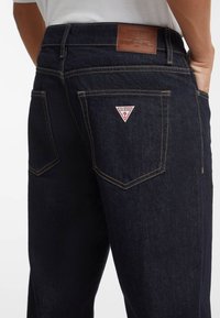 Jeans di denim scuro con vestibilità dritta. Presentano cuciture dorate, una patta in pelle con il logo e un'etichetta triangolare rossa del brand sulla tasca posteriore.