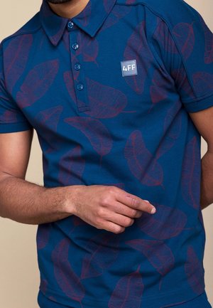 Mand iført blå poloshirt med lilla bladmotiver og et lille 4FF-logo på brystet, arm bøjet med hånden nær taljen.