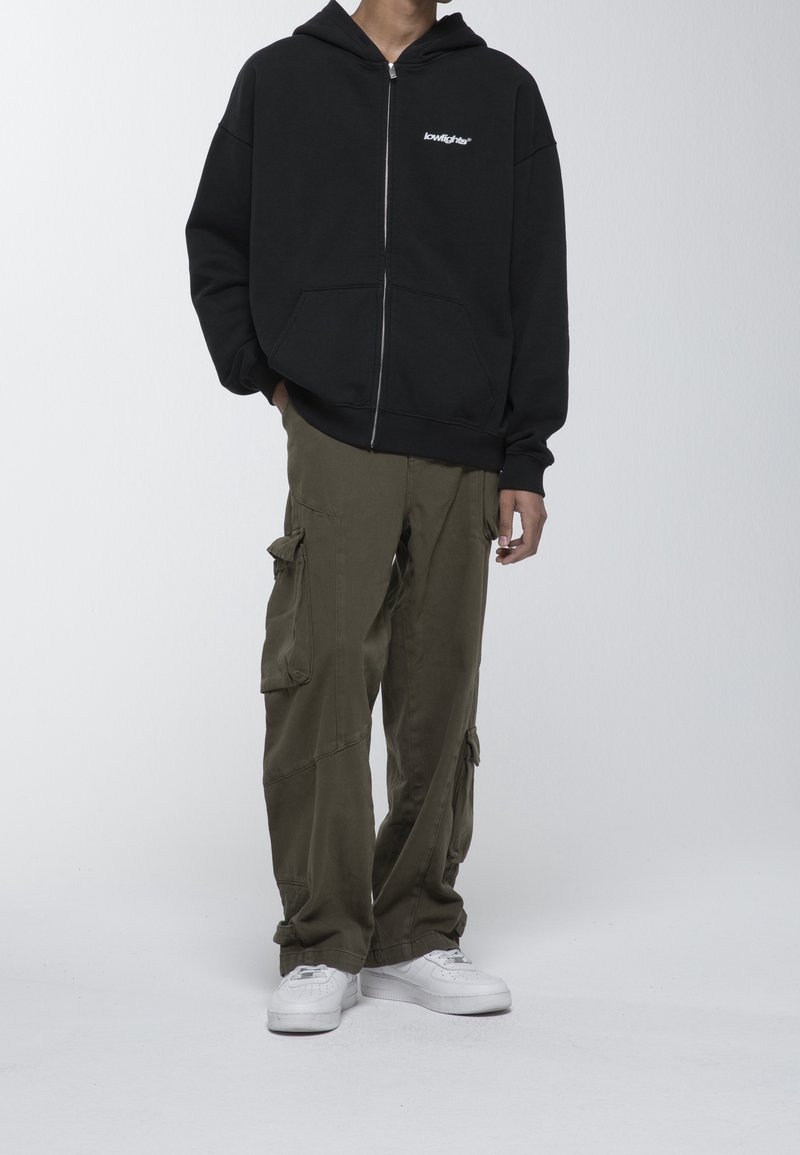 Personne portant un sweat à capuche noir zippé avec le texte "lowlights", un pantalon cargo vert olive et des baskets blanches sur un fond uni.