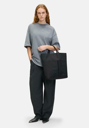 Schwarze Tragetasche aus strapazierfähigem Material, mit doppelten Griffen und einer Vordertasche. Getragen zu einem lockeren grauen T-Shirt und weiten schwarzen Hosen.