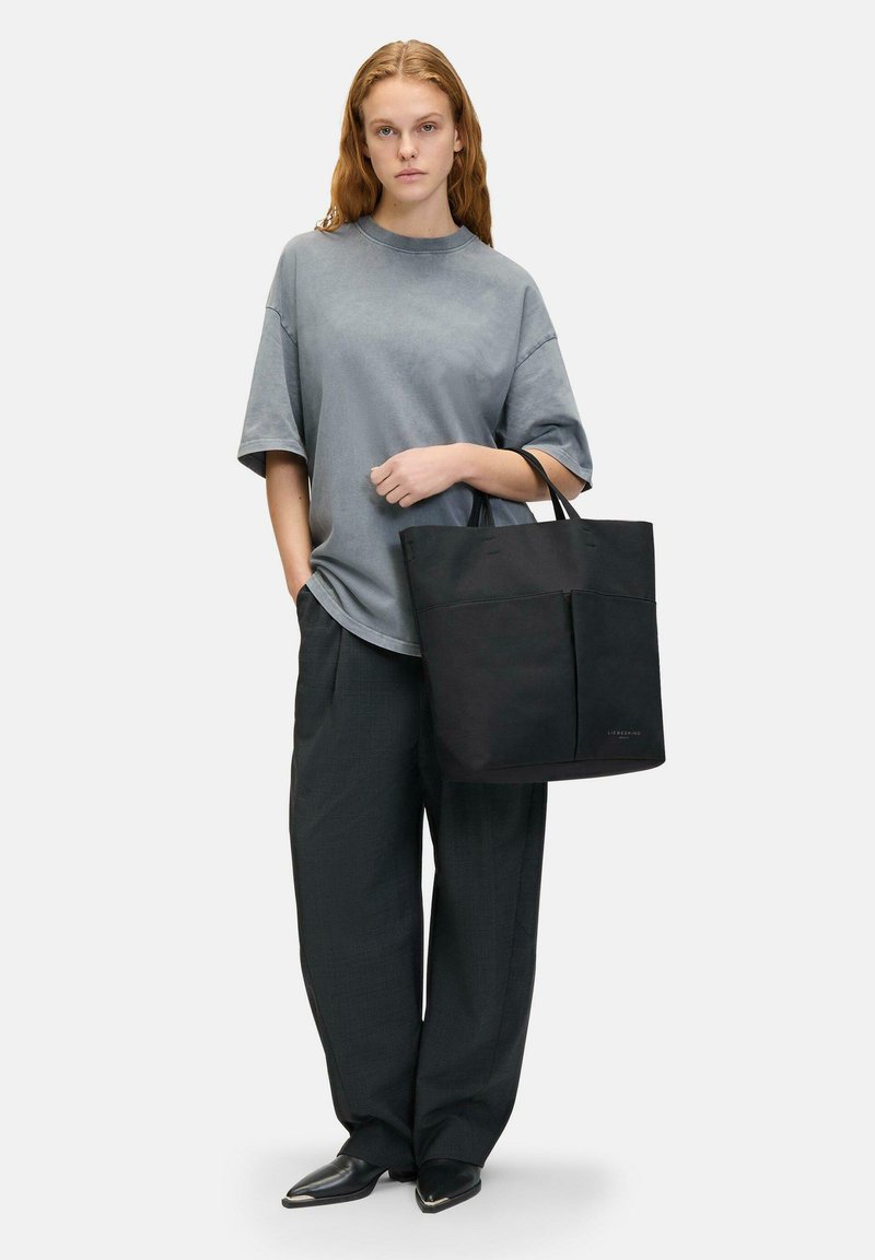 Borsa a tracolla nera realizzata in materiale resistente, con doppie maniglie e una tasca frontale. Indossata con una t-shirt grigia oversize e pantaloni neri larghi.