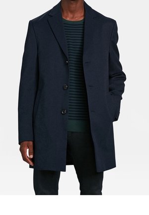 Homme portant un manteau bleu marine foncé à trois boutons sur un pull rayé vert foncé et noir, une main dans la poche du manteau.