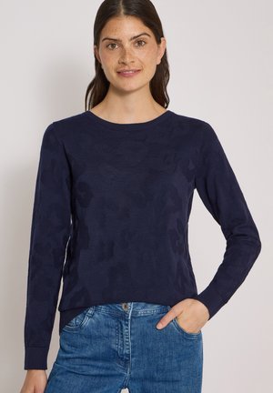 Femme aux longs cheveux foncés portant un pull bleu marine à motifs et un jean bleu, une main dans la poche, souriant légèrement.