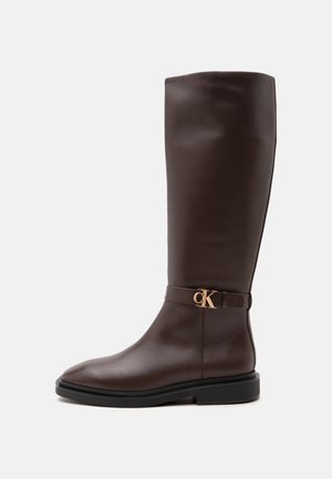 Bota de cuero marrón oscuro hasta la rodilla con suela negra y pequeño detalle de logo dorado "CK" en la correa cerca del tobillo, mostrado de lado.