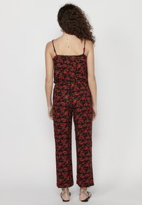 Floral jumpsuit met een zwarte achtergrond en rode en groene bloemen, dunne verstelbare schouderbanden, een losse pasvorm en wijde broekspijpen.