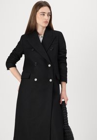 Cappotto nero doppiopetto con grandi bottoni, a rever e tasche laterali. Tessuto liscio con una vestibilità su misura e lunghezza completa.