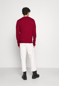 Maglione rosso a maglia con maniche lunghe, colletto rotondo e bordo a coste. Abbinato a pantaloni bianchi e stivali neri con lacci. Sfondo semplice.
