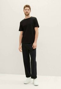 Jovem de pé, vestindo uma t-shirt preta simples, calças pretas com punhos e ténis brancos, em frente a um fundo neutro.