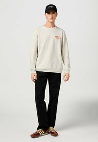 Felpa grigia chiara con grafica arancione, pantaloni neri e scarpe da ginnastica marroni con dettagli gialli. Cappellino nero con logo bianco. Abbigliamento casual.