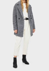 Gray, textured wool-blend coat with a double-breasted design, large collar, and black buttons, worn over a white sweater and cream pants. Black ankle boots.
