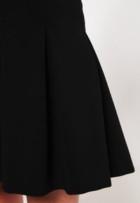 Musta A-linjainen hame, joka on valmistettu sileästä kankaasta ja jossa on laskoksia lisävolyymia antamassa sekä istuva vyötärönsuunnittelu, joka luo puhtaan ja minimalistisen ilmeen.