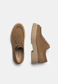 Scarpe in suede marroni con punta arrotondata, lacci elasticizzati e una suola in gomma spessa e rigata, con un sottile dettaglio di cucitura lungo il bordo.