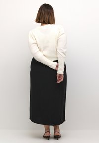 Crème-kleurige trui met poffende mouwen en textuurmanchetten, gedragen met een lange zwarte rok, gezien vanaf de achterkant, met zwarte haksandalen.