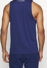 Uomo che indossa una maglietta atletica senza maniche blu navy di Tommy Sport e pantaloni atletici coordinati blu navy, visti da dietro.