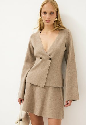 Mujer con top beige de punto con estilo envolvente y mangas largas, y falda a juego, sosteniendo un bolso de color claro, de pie contra una pared blanca.