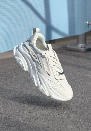 Weißer Steve Madden Sneaker mit dicker Sohle und Mesh-Details, schwebend in der Luft über Asphaltboden nahe eines blauen Zauns und eines Metallpfostens.
