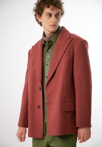 Czerwony płaszcz z wełny z klapami, z podwójnym zapięciem na guziki i dwiema przednimi kieszeniami. Noszony nad zieloną koszulą i jasnoróżowym golfem.