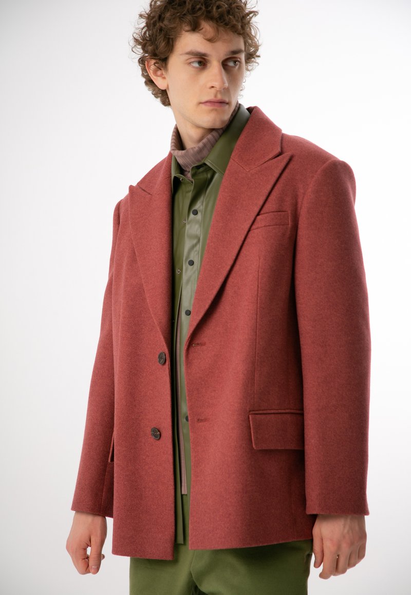 Czerwony płaszcz z wełny z klapami, z podwójnym zapięciem na guziki i dwiema przednimi kieszeniami. Noszony nad zieloną koszulą i jasnoróżowym golfem.