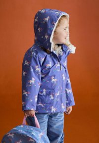 Lila wasserdichte Jacke mit Einhornmuster, weiße Fleece-futterte Kapuze und zwei Fronttaschen. Kind hält einen blauen gemusterten Rucksack.